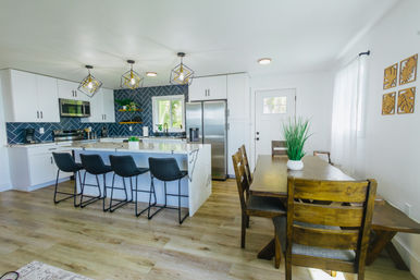 Sunlit open-concept modern kitchen and dining area with marble island and four black bar stools, navy herringbone tile backsplash, stainless-steel appliances, wooden dining table, pendant lights and light wood floors.