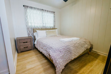 Cozy bright bedroom with textured pale-pink bedspread, white pillows and folded towels on the bed, wooden nightstand, patterned sheer curtains and light wood plank flooring.