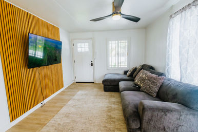 Bright modern living room with gray sectional sofa and patterned pillows, wood-slat accent wall with wall-mounted TV, ceiling fan, area rug, and front door with window.
