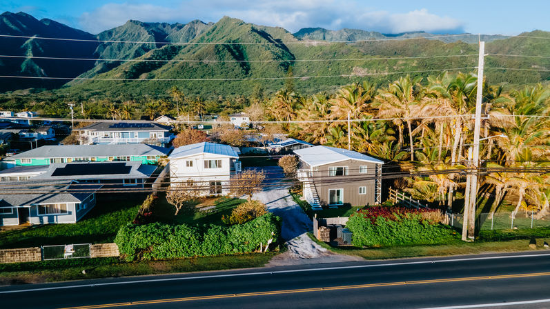 Aerial view of a sunny tropical neighborhood with palm trees, small coastal homes along a two-lane road and lush green mountains rising in the background.