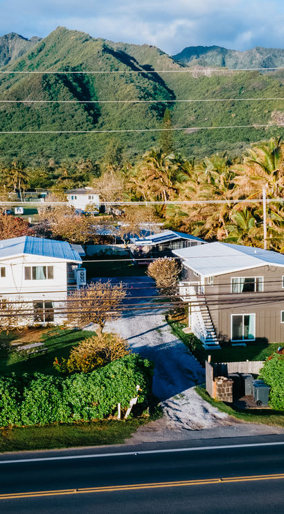 Aerial view of a sunny tropical neighborhood with palm trees, small coastal homes along a two-lane road and lush green mountains rising in the background.