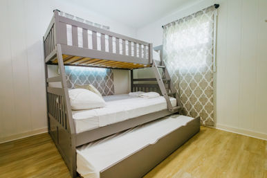 Cozy guest bedroom with gray wooden bunk bed, ladder and pull-out trundle, white linens, patterned beige curtains and light hardwood floor.