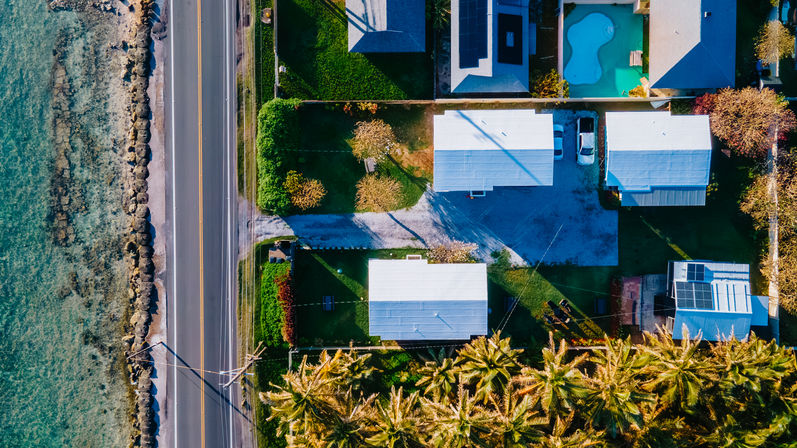 Aerial shot of a tropical coastline: a two-lane road beside a rocky seawall and clear shallow water, adjacent to white-roofed houses with driveways, a turquoise backyard pool and swaying palm trees casting long shadows.