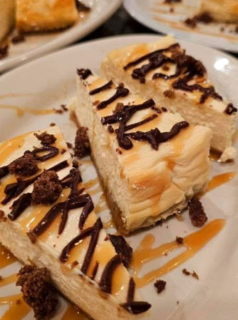 Close-up of creamy cheesecake bars with graham crust on a white plate, drizzled with chocolate and caramel and sprinkled with chocolate crumble — a decadent dessert.