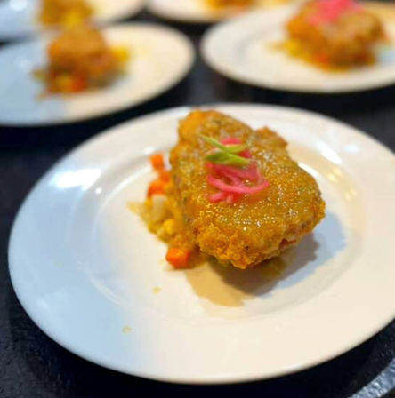 Crunchy golden-brown breaded cutlet topped with bright pickled red onions and a green garnish, served over diced vegetables on a white plate as a plated appetizer.