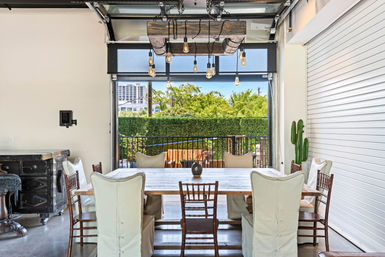 Modern industrial indoor-outdoor dining area with farmhouse table and mixed cushioned and wooden chairs, hanging Edison-bulb chandelier, open glass roll-up door to a sunny balcony, green hedge and distant urban buildings