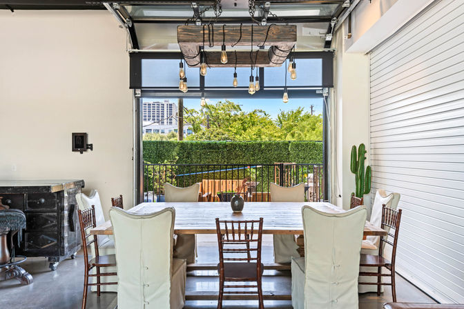 Modern industrial indoor-outdoor dining area with farmhouse table and mixed cushioned and wooden chairs, hanging Edison-bulb chandelier, open glass roll-up door to a sunny balcony, green hedge and distant urban buildings