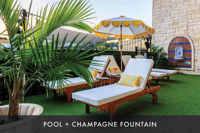 Sunny outdoor poolside lounge with white cushioned wooden sunbeds, yellow-striped pillows and scalloped yellow umbrella on green turf, potted palms and a decorative tiled champagne fountain in a stone wall.