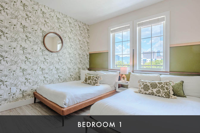 Sunny modern bedroom with two platform beds, botanical wallpaper, round wall mirror, green accent panels, bedside lamp, and city view through large windows.