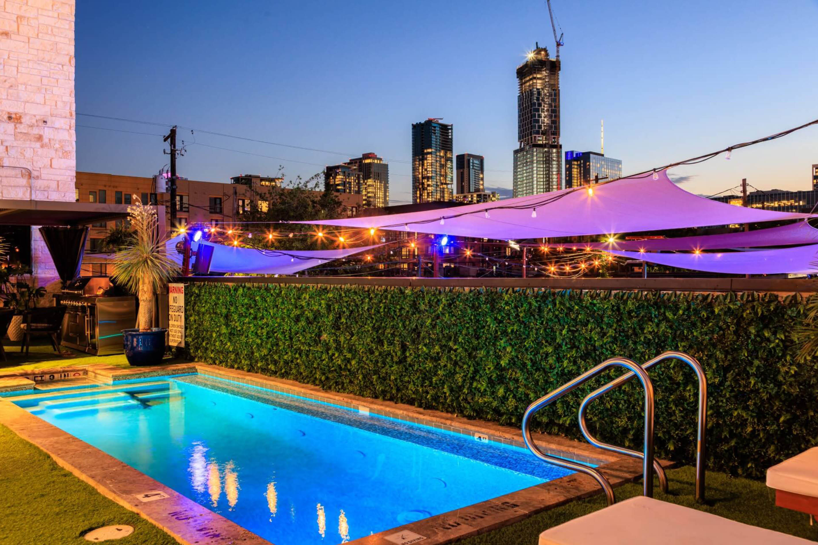 Rooftop illuminated pool at dusk with glowing blue water, string lights and purple shade sails, overlooking a modern downtown skyline.