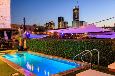 Rooftop illuminated pool at dusk with glowing blue water, string lights and purple shade sails, overlooking a modern downtown skyline.