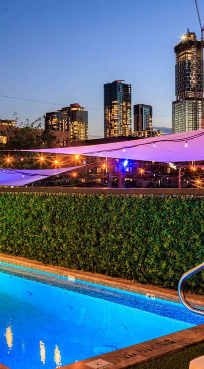 Rooftop illuminated pool at dusk with glowing blue water, string lights and purple shade sails, overlooking a modern downtown skyline.