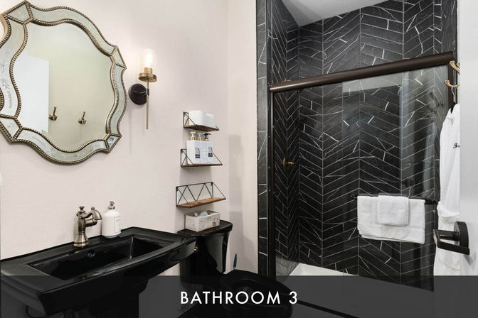 Stylish modern bathroom interior with black pedestal sink and toilet, glass shower with dark herringbone tile and bronze hardware, ornate beveled mirror, brass wall sconce, floating shelves with toiletries and white towels.