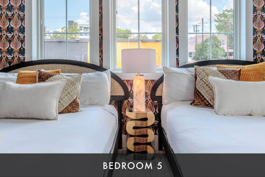 Bright twin guest room with two white-made beds and woven headboards, patterned wallpaper, mustard and textured throw pillows, sculptural wooden nightstand topped by a marble lamp, and large windows with an urban city view.