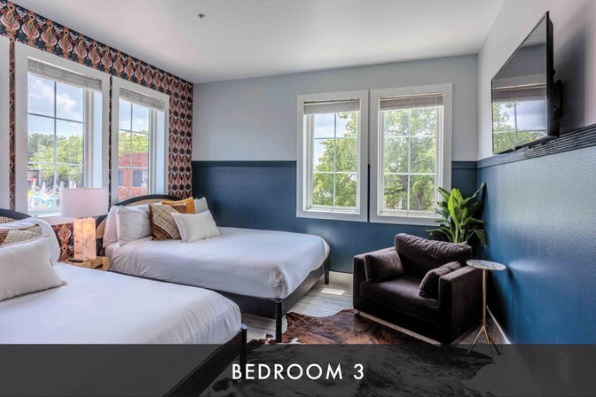 Bright modern guest room with two beds, navy half-wall and patterned accent wallpaper, large windows with tree and brick building views, plush armchair and wall-mounted TV.