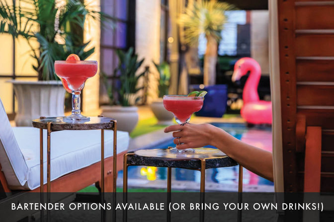 Poolside scene: two pink citrus-garnished cocktails on small side tables beside a lounge chair, a hand reaching for one, tropical potted plants and a pink flamingo pool float in the blurred background — relaxed outdoor pool lounge vibe.