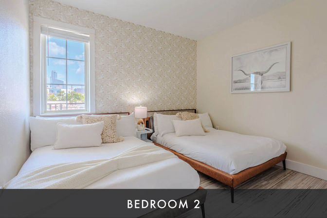 Bright modern bedroom with two beds, neutral linens and patterned accent wall, cozy pillows and bedside lamp, with downtown city skyline visible through the window.