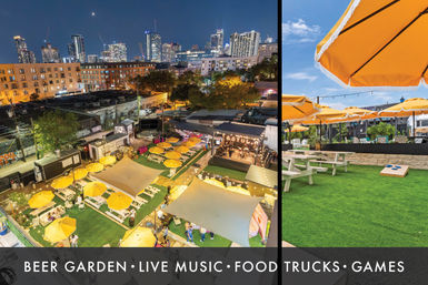 Rooftop beer garden in a downtown skyline setting with yellow umbrellas over picnic tables on green turf, food trucks, a small stage for live music, outdoor games and string lights at dusk.