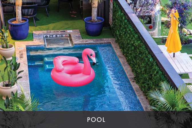 Bright pink inflatable flamingo floating in a small rectangular outdoor pool with clear blue water, surrounded by potted plants, turf patio seating, a green hedge and a yellow umbrella