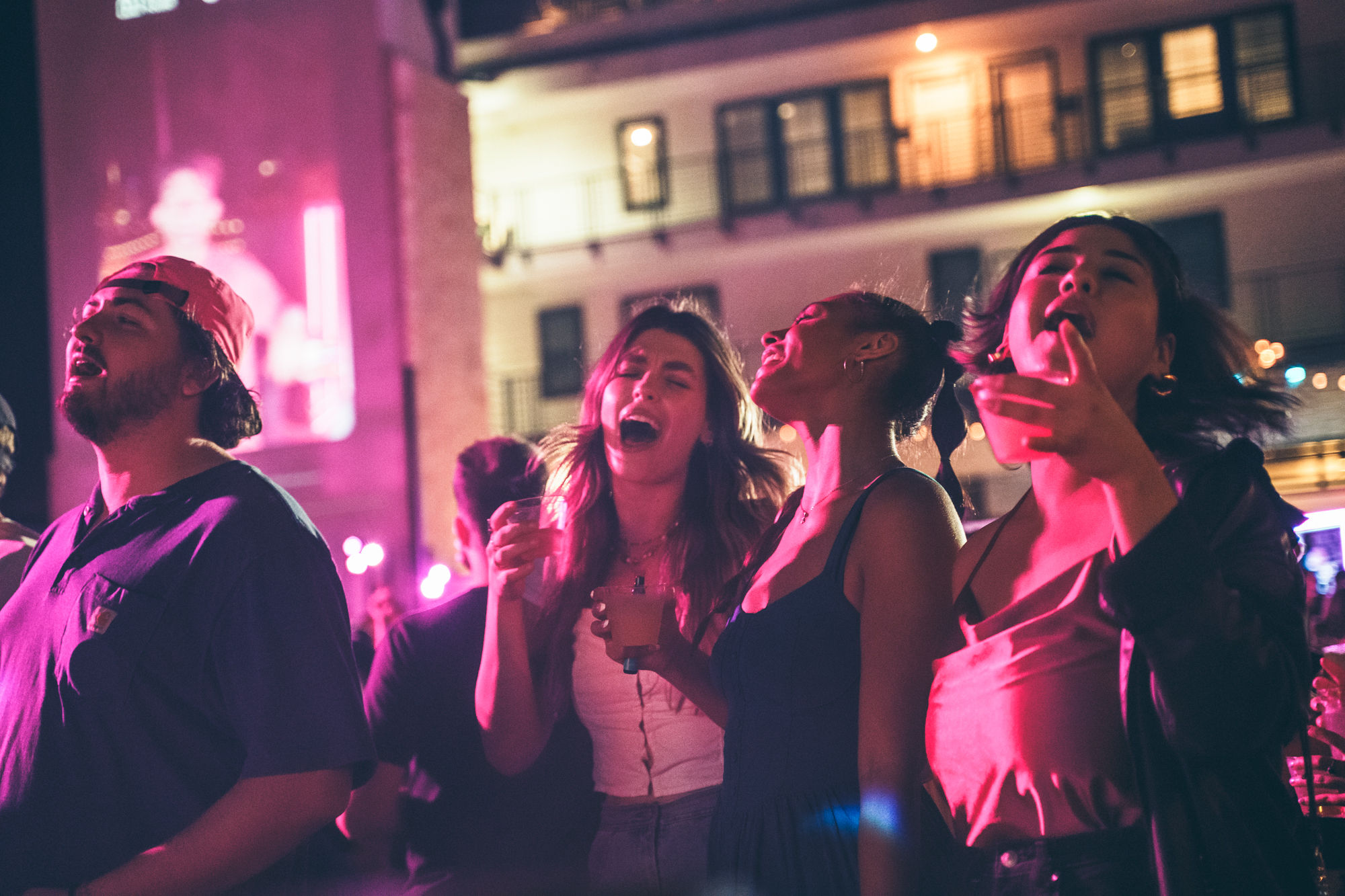 Group of friends singing and holding drinks at a neon-lit outdoor nightlife party in a downtown courtyard