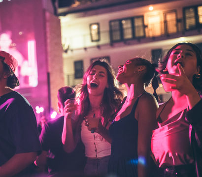 Group of friends singing and holding drinks at a neon-lit outdoor nightlife party in a downtown courtyard