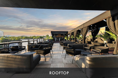 Rooftop lounge with modern sofas, black chairs and shaded cabanas on a wooden deck, potted palms and tables overlooking a city skyline at sunset