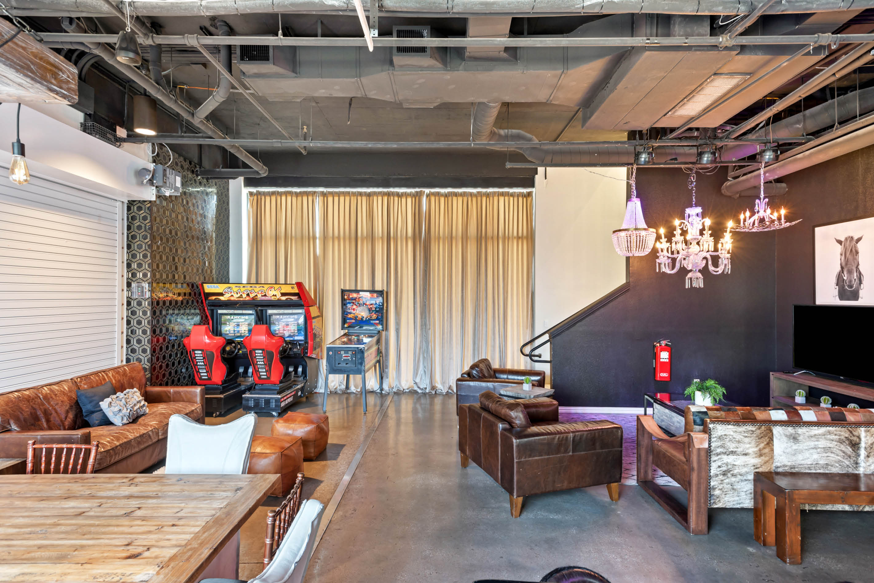 Industrial loft-style game lounge with red arcade racing cabinets and pinball machine, leather sofas, rustic wooden table, chandeliers, and exposed ductwork.
