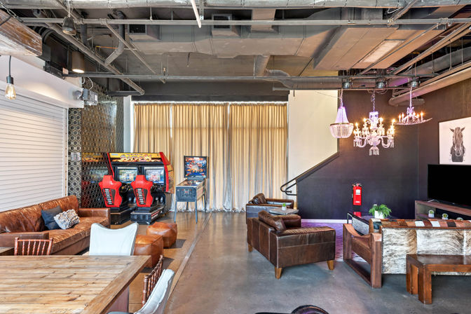 Industrial loft-style game lounge with red arcade racing cabinets and pinball machine, leather sofas, rustic wooden table, chandeliers, and exposed ductwork.