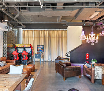 Industrial loft-style game lounge with red arcade racing cabinets and pinball machine, leather sofas, rustic wooden table, chandeliers, and exposed ductwork.