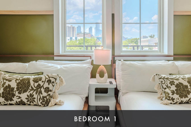 Cozy twin beds with crisp white linens and floral tassel pillows, shared nightstand and lamp, window framing a sunny downtown city skyline view.