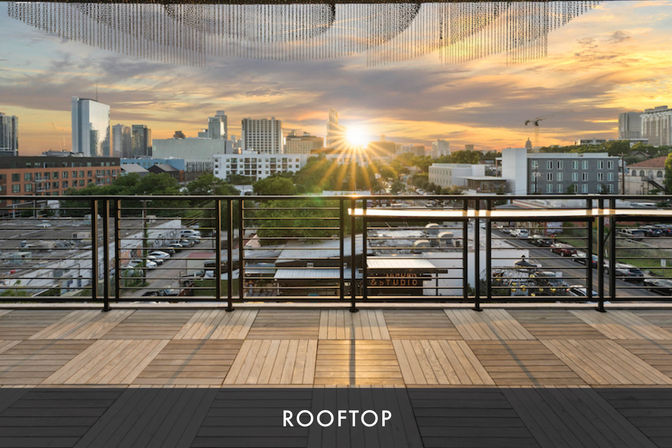 Rooftop terrace with patterned wooden deck and metal railing overlooking a downtown city skyline at golden sunset, sunburst glinting between buildings.