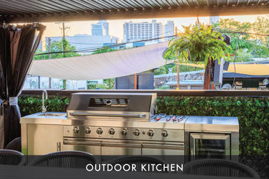 Stainless-steel outdoor kitchen with large gas grill, side sink, prep counter and fridge on a shaded urban rooftop patio with a hanging fern and city skyline