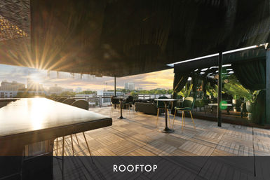 Sunset rooftop terrace with sleek bar, high-top tables and plush green chairs beside potted palms, open-air lounge overlooking an urban skyline