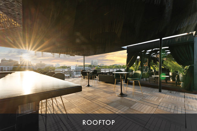 Sunset rooftop terrace with sleek bar, high-top tables and plush green chairs beside potted palms, open-air lounge overlooking an urban skyline