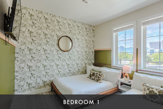 Bright urban bedroom with botanical wallpaper, round mirror, wall-mounted TV, two white-made beds with patterned throw pillows, bedside table with lamp, and large windows showing a city view.