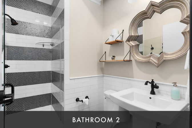 Chic small bathroom with walk-in shower featuring black hex-tile stripes and white subway tile, white pedestal sink, ornate quatrefoil mirror, wooden floating shelves with toilet paper and a plant, and matte black fixtures.