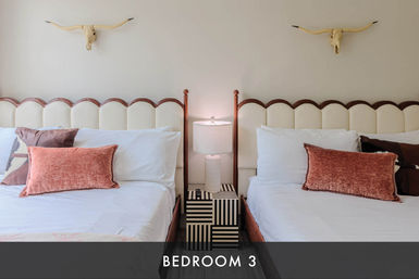 Bright guest bedroom with two twin beds, scalloped cream headboards, coral velvet throw pillows, striped black-and-white nightstand and lamp between beds, and decorative longhorn skulls on the wall.
