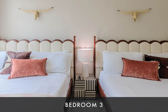 Bright guest bedroom with two twin beds, scalloped cream headboards, coral velvet throw pillows, striped black-and-white nightstand and lamp between beds, and decorative longhorn skulls on the wall.