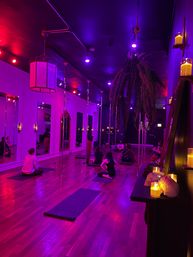 Purple-lit urban fitness studio interior with hardwood floor, mirrored wall, vertical poles, hanging fern and candles, people seated on yoga mats
