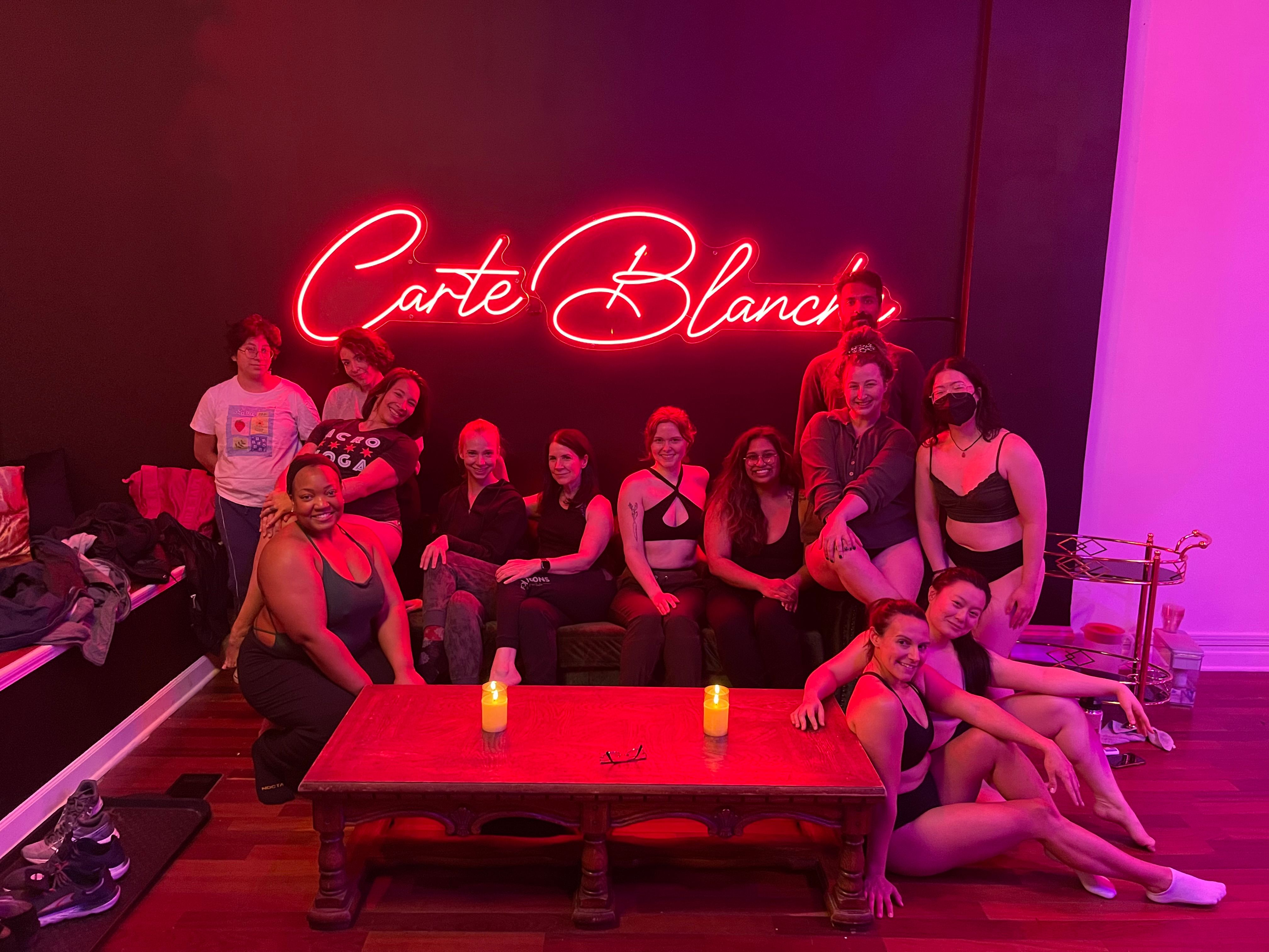 Diverse group of adults in workout clothes smiling and posing in a pink neon-lit studio lounge around a wooden coffee table with lit candles, post-class relaxed vibe.