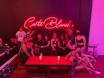 Diverse group of adults in workout clothes smiling and posing in a pink neon-lit studio lounge around a wooden coffee table with lit candles, post-class relaxed vibe.