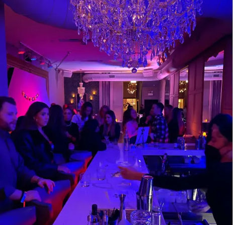 Bustling cocktail lounge interior at night — patrons seated along a white bar under a sparkling crystal chandelier, purple-blue mood lighting, bartender preparing drinks for an intimate urban nightlife crowd.