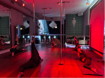 Red-lit pole fitness studio with students stretching on mats around vertical poles, mirrored walls, wooden floor, chandeliers and bar seating in a stylish urban workout space