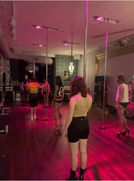 Group pole dancing class in a dim studio with pink uplighting, chrome poles, hardwood floors and students practicing in high heels.
