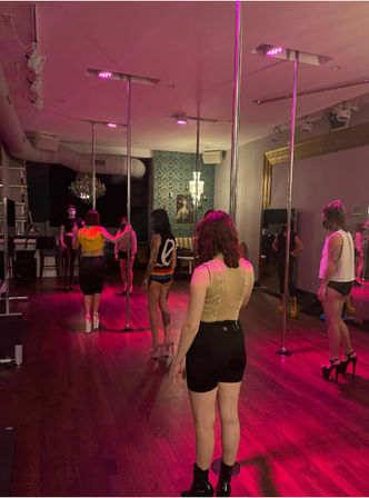 Group pole dancing class in a dim studio with pink uplighting, chrome poles, hardwood floors and students practicing in high heels.