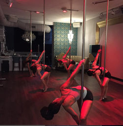 Pole fitness class in a dim studio: a group of women practicing dynamic pole-dance moves under red lighting on a hardwood floor with a chandelier and patterned wallpaper.