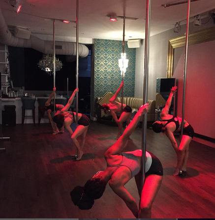 Pole fitness class in a dim studio: a group of women practicing dynamic pole-dance moves under red lighting on a hardwood floor with a chandelier and patterned wallpaper.