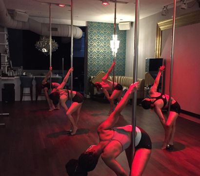 Pole fitness class in a dim studio: a group of women practicing dynamic pole-dance moves under red lighting on a hardwood floor with a chandelier and patterned wallpaper.
