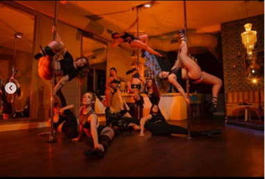 Group pole dance fitness class inside a studio, several dancers performing on chrome poles and on the floor under warm amber lighting with mirrored wall and lounge seating