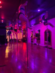 Energetic pole fitness class in a pink-purple lit studio — participants practicing spins and climbs on chrome poles beside mirrors over a polished hardwood floor.